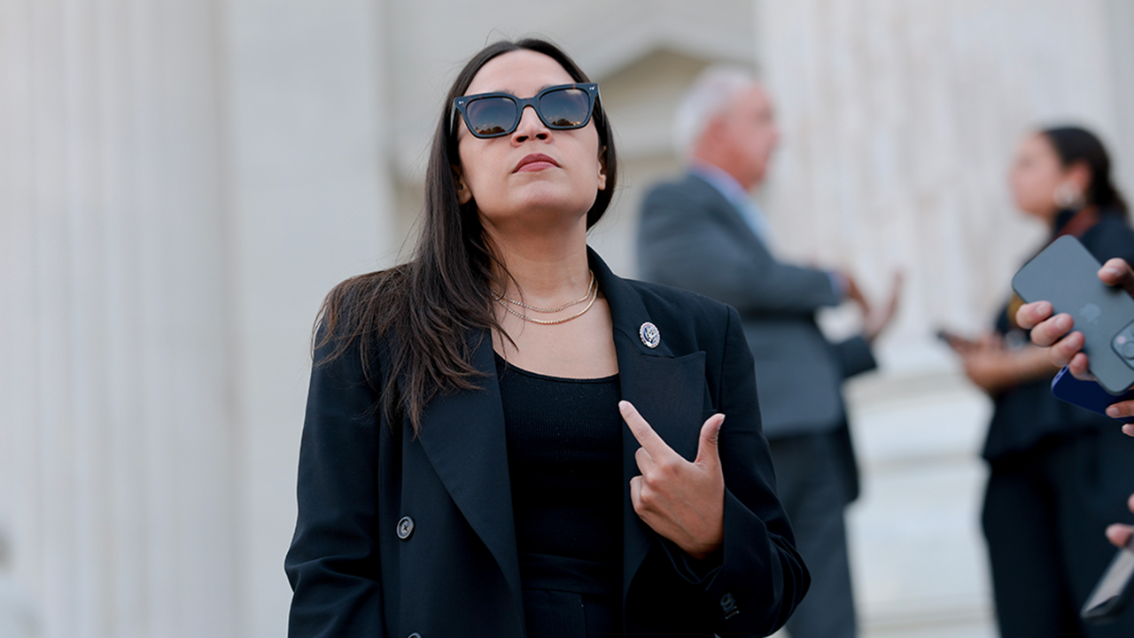 A woman in a black outfit and sunglasses stands outside, pointing at herself, with people and white columns blurred in the background.