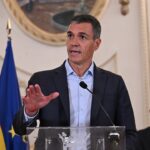 A man in a suit speaks at a podium with flags behind him, including the Spanish and EU flags, inside an ornate room. Another man is seated in the background, and a decorative round frame hangs on the wall.