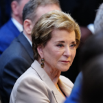 A woman with short light brown hair and a beige blazer sits among other people in a formal setting, looking slightly to the side. The background is out of focus, highlighting her face.