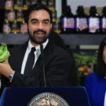 A man in a suit holds up a bunch of green bananas while speaking at a podium. A woman in a blue jacket stands beside him. Grocery products are visible on shelves in the background.