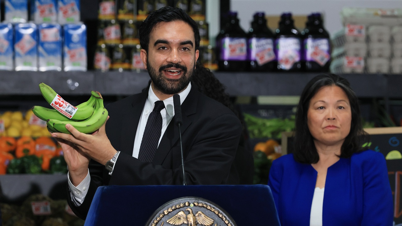 A man in a suit holds up a bunch of green bananas while speaking at a podium. A woman in a blue jacket stands beside him. Grocery products are visible on shelves in the background.