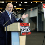 Steve Hilton at a podium with a Migrant Child Abuse Camps sign, while a woman sits nearby. Beside him is a large Wanted poster featuring a mans photo and the name Xavier Becerra.
