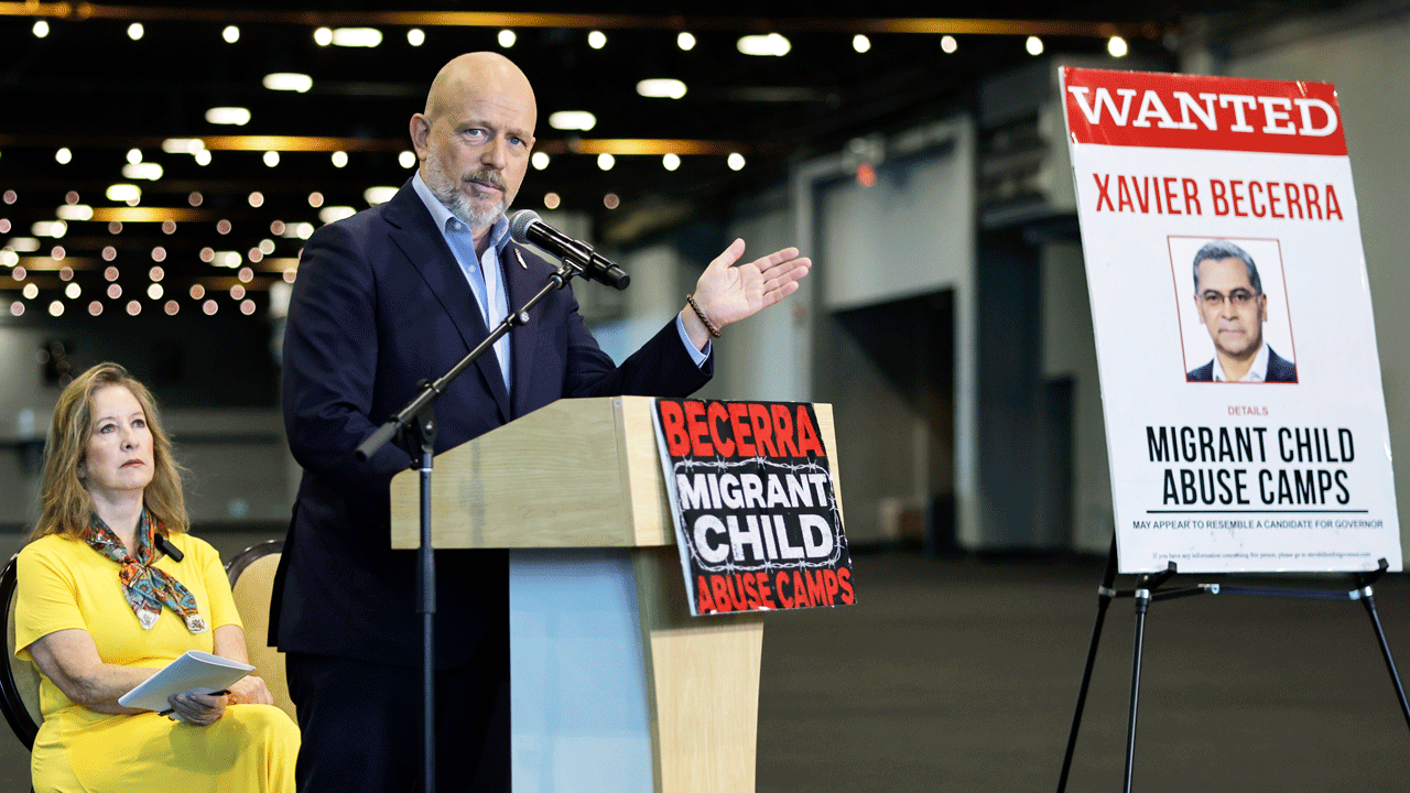 Steve Hilton at a podium with a Migrant Child Abuse Camps sign, while a woman sits nearby. Beside him is a large Wanted poster featuring a mans photo and the name Xavier Becerra.