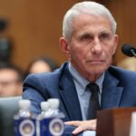 An older man in a blue suit sits at a panel during a hearing. Microphones and two water bottles are in front of him, with blurred attendees seated in the background.