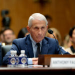 Dr. Anthony Fauci sits at a table with a somber expression on his face while wearing a dark suit during a congressional hearing.