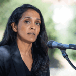 A woman with long dark hair, wearing a black blazer, stands outdoors speaking into a microphone. Greenery appears blurred in the background.