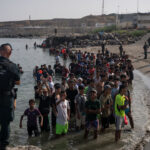 A group of men stand in shallow water while uniformed officers watch them. Buildings and a fence are visible in the background.