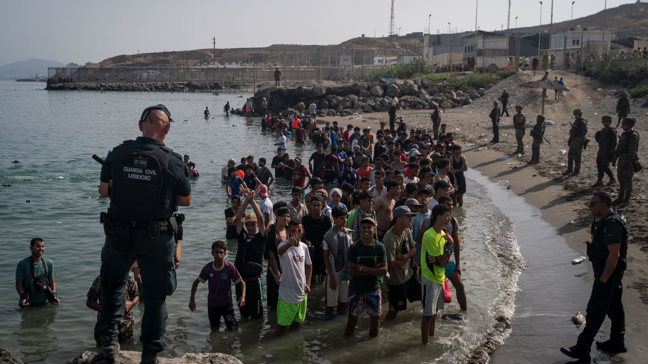 A group of men stand in shallow water while uniformed officers watch them. Buildings and a fence are visible in the background.