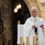 A clergy member in white and gold robes stands at a lectern, holding papers and speaking in a decorated church. Three lit candles and a flower arrangement are visible nearby.