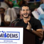 A man speaks enthusiastically at a podium labeled MIDEMS with people in the background holding signs at a public event or rally.