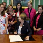 A woman sits at a desk signing a document, surrounded by a group of women standing and smiling in a formal room.