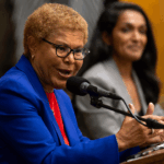 Mayor Karen Bass, wearing a blue blazer, speaks into a microphone while her hands are at a podium.