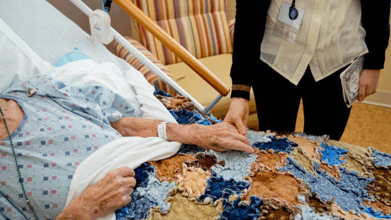 A hospice patient is in bed with her hand being held by a hospice caregiver.