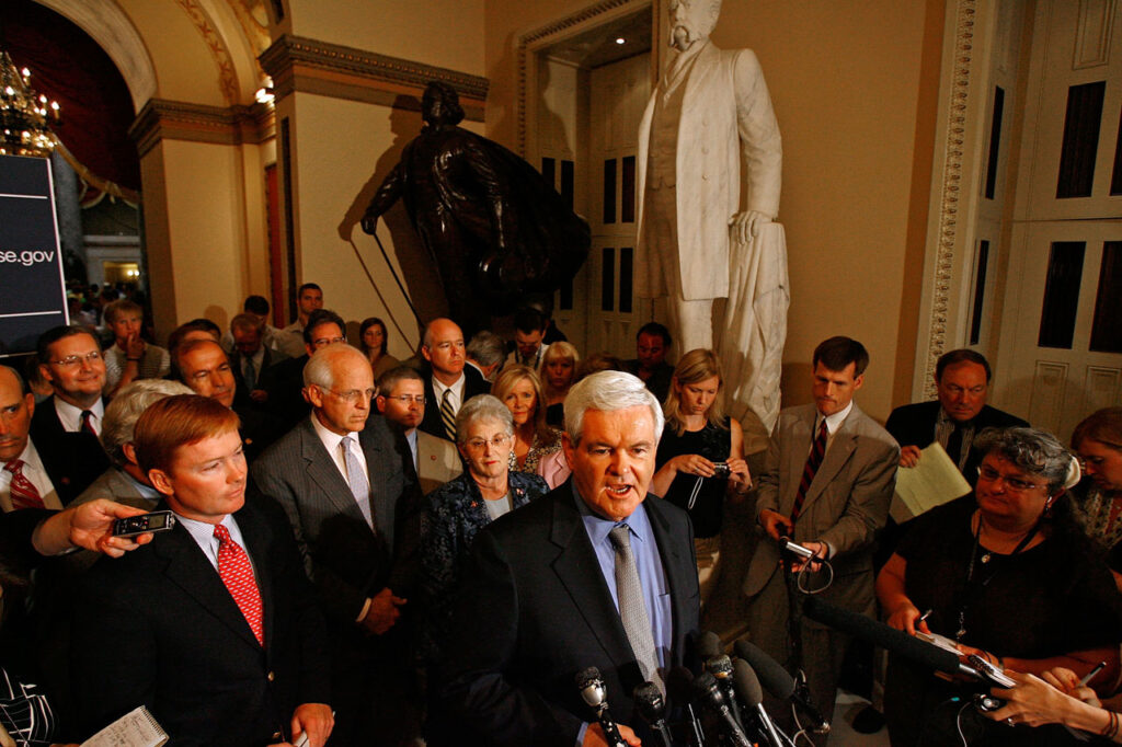 A man speaks to reporters in a crowded hallway lined with statues. Microphones and cameras surround him, while people behind him listen, take notes, and watch attentively. The setting appears to be a government building.