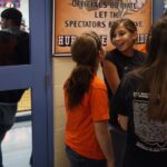 A group of girls in orange and black shirts stand and talk near a gym entrance.