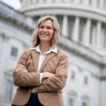 A woman in a tan blazer stands confidently with arms crossed in front of a government building with columns and a dome, smiling at the camera.