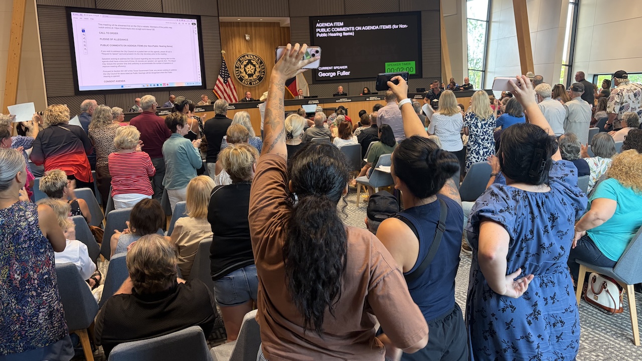 A large crowd attends a public meeting in a modern room; many people are standing, some with raised fists, facing officials seated at a long front desk with digital displays and American flags.