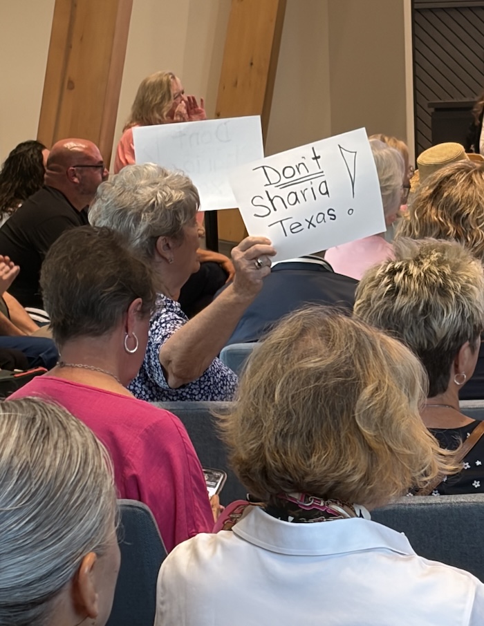 McKinney City Council Unanimously Approves Mosque After Hours of Public Opposition 1 A woman in a crowded room holds up a sign that reads, Dont Sharia Texas! Several people are seated around her, facing forward.