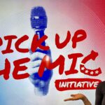 A woman smiles and gestures toward a screen displaying “PICK UP THE MIC INITIATIVE” in bold red letters with a graphic of a hand holding a microphone in the background.