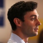 Senator Jon Ossoff appears with short dark hair, wearing a light blue collared shirt. He is shown in profile, speaking indoors with blurred people and signs in the background.