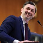 Senator Jon Ossoff appears in a navy suit and red tie. He smiles while seated at a desk with a microphone, in a formal wood-paneled room.