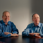 Steven Brill and L. Gordon Crovitz sit at a brown table in front of a gray wall.