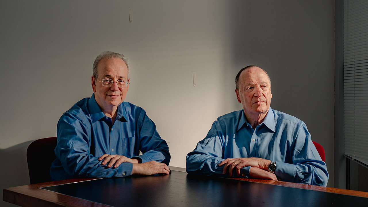 Steven Brill and L. Gordon Crovitz sit at a brown table in front of a gray wall.