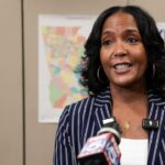 Keisha Lance Bottoms appears in a striped blazer and speaks at a press event with microphones in front of her; campaign signs reading Keisha Lance Bottoms Governor are visible on the wall behind her.