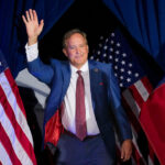 Ken Paxton in blue suit with red tie waves as he comes on a stage in front of American flags.