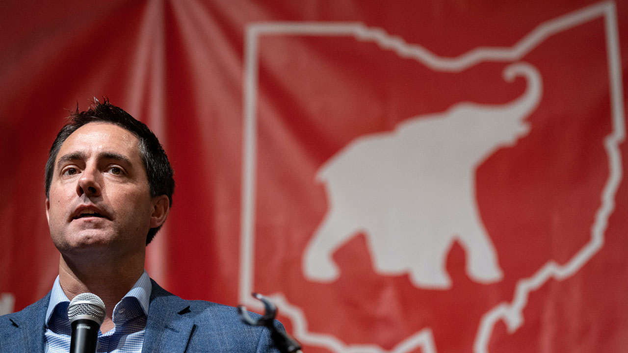 Ohio Secretary of State Frank LaRose speaks in front of a red banner with an elephant inside a map of Ohio.