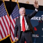 Mike Collins stands and waves in a suit and red tie. He appears on stage in front of two U.S. flags.