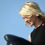 Natalie Harp, a top Trump aide, appears with blonde hair, wearing a sleeveless black top and backpack. She looks down and smiles against a clear blue sky.