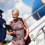 Trump Aide Natalie Harp holds a folder and exits an airplane, flanked by two uniformed Air Force members standing guard. The sky is blue with clouds in the background.