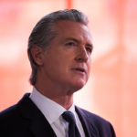 Gavin Newsom, stands in front of a softly blurred red background, looking slightly upward with a serious expression.