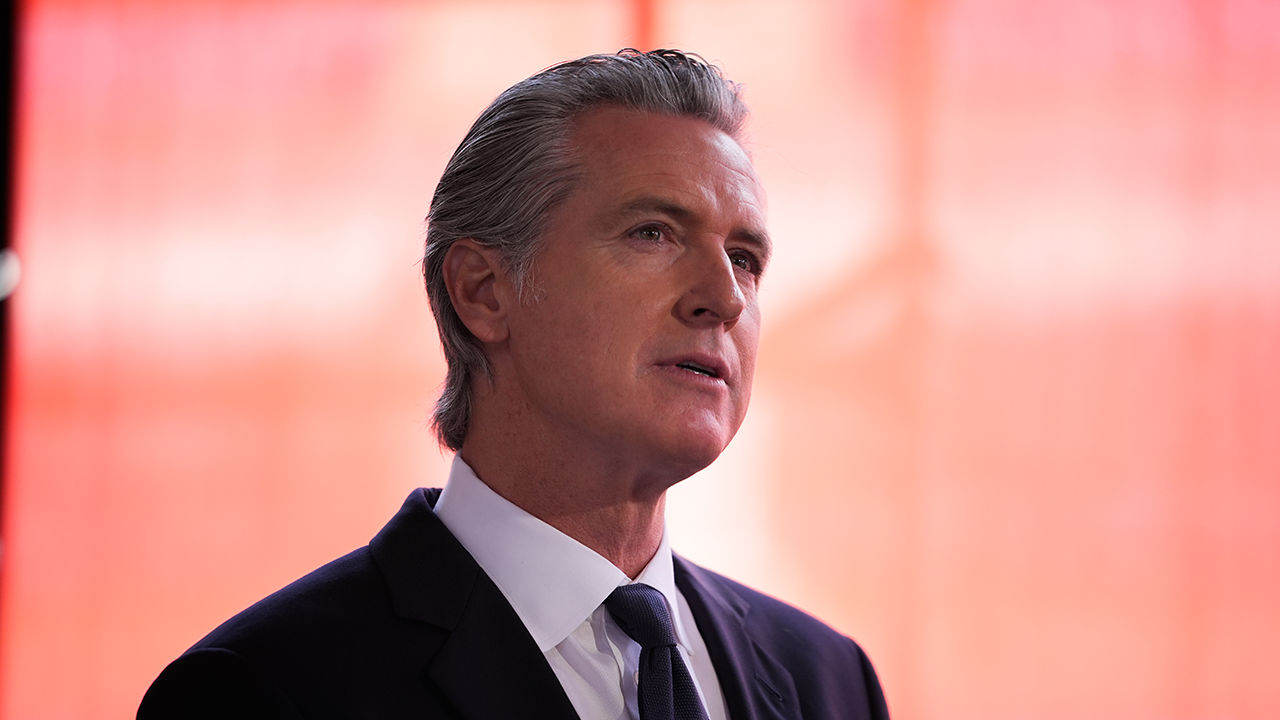 Gavin Newsom, stands in front of a softly blurred red background, looking slightly upward with a serious expression.