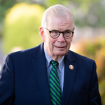 Rep. Tim Walberg standing outdoors amidst blurred greenery and sunlight, his thoughtful expression suggesting a psychological depth to his character.