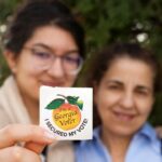 Two women are pictured, while one of them holds a Georgia voter sticker in front of a tree.