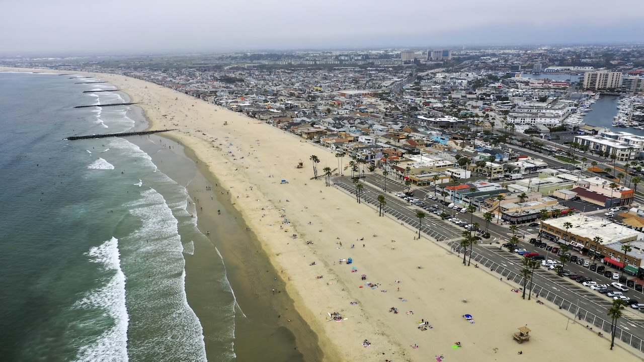Aerial view of a long sandy beach with waves, scattered beachgoers, palm trees, parked cars, and a densely built coastal city under a cloudy sky.