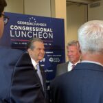 Rick Jackson and Gov. Brian Kemp appear in suits and speak in front of a sign that reads Georgia Chamber Congressional Luncheon while microphones are held toward them for an interview.