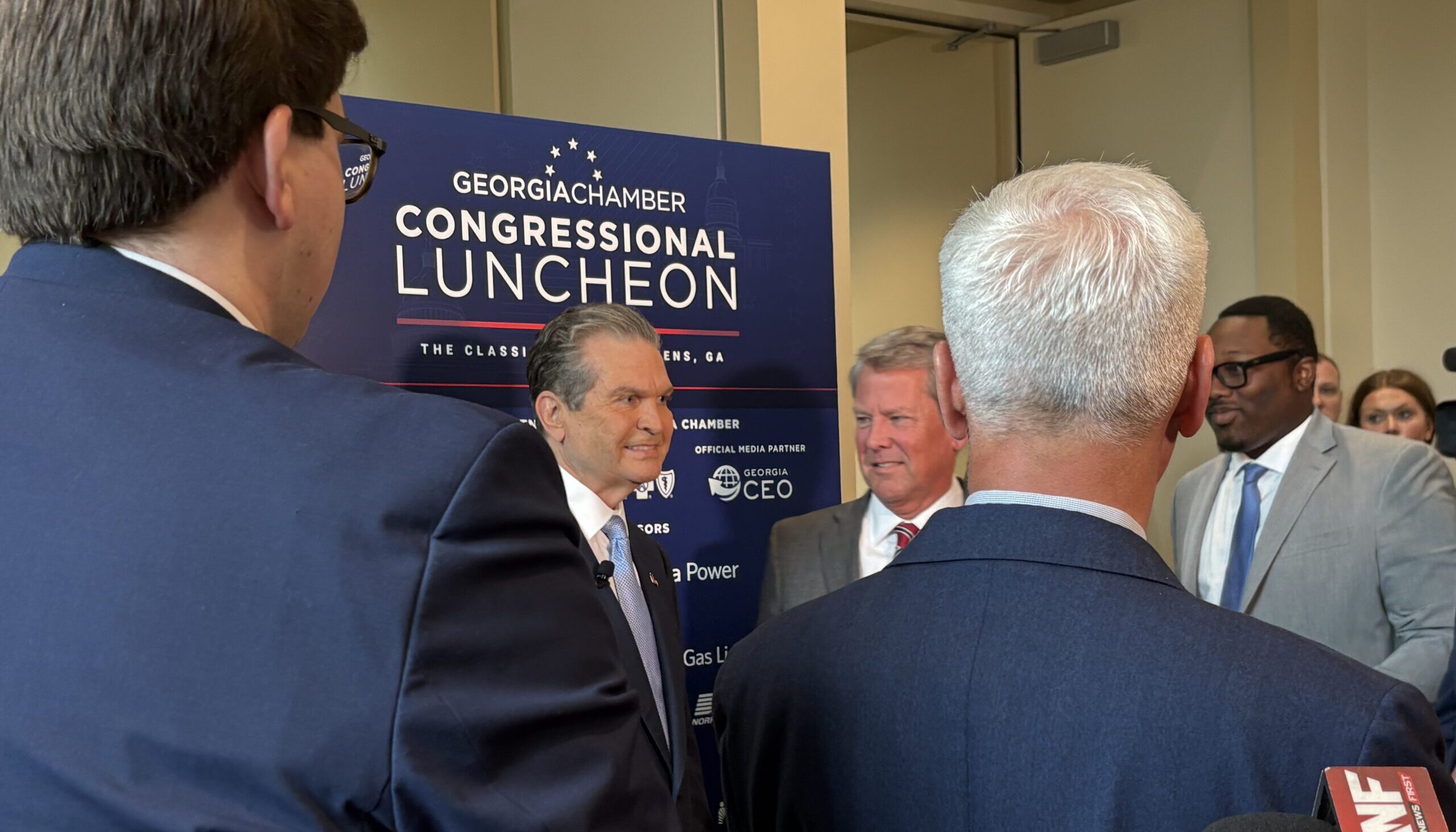 Rick Jackson and Gov. Brian Kemp appear in suits and speak in front of a sign that reads Georgia Chamber Congressional Luncheon while microphones are held toward them for an interview.