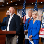 A group of people in business attire stand at a podium with U.S. flags behind them. A sign reads, “Stalled Out Senate: Pass the Save America Act!” The speaker gestures while addressing the audience.