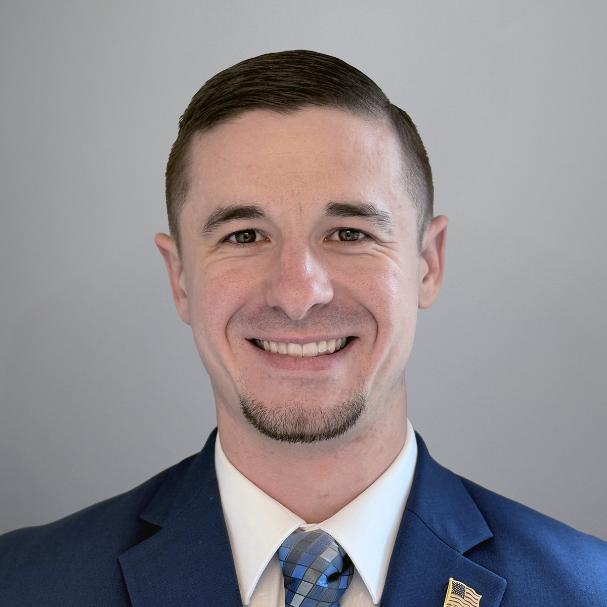 Smiling man with short brown hair wearing a blue suit, white shirt, and plaid blue tie, standing against a light gray background.