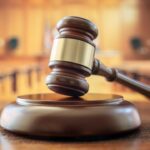 A wooden gavel rests on a sound block in an empty courtroom, symbolizing justice in cases of fraud, with the blurred judge’s bench and seating visible in the background.
