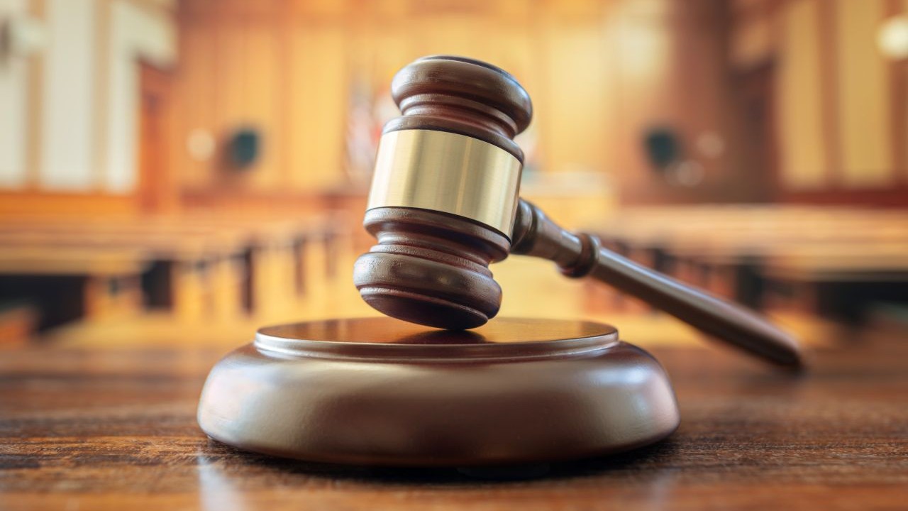 A wooden gavel rests on a sound block in an empty courtroom, symbolizing justice in cases of fraud, with the blurred judge’s bench and seating visible in the background.