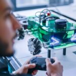 A Siemens trainee holds a game controller, operating a green robotic vehicle with LED lights and mecanum wheels on a table in a modern lab.