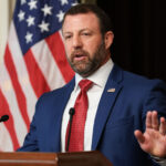 A bearded man in a blue suit speaks at a podium, gesturing with one hand. An American flag is visible in the background.