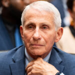 An older man with short white hair in a blue suit sits thoughtfully with his hand on his chin at what appears to be a formal hearing, with blurred people in the background.