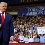 President Trump appears in a blue suit and red tie. He stands in front of a large, cheering crowd holding signs at an indoor event. A banner behind him reads POWERING THE AMERICAN DREAM.