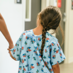 A young girl in a hospital gown with a braid walks hand-in-hand with a healthcare worker down a hallway. (Getty Images)