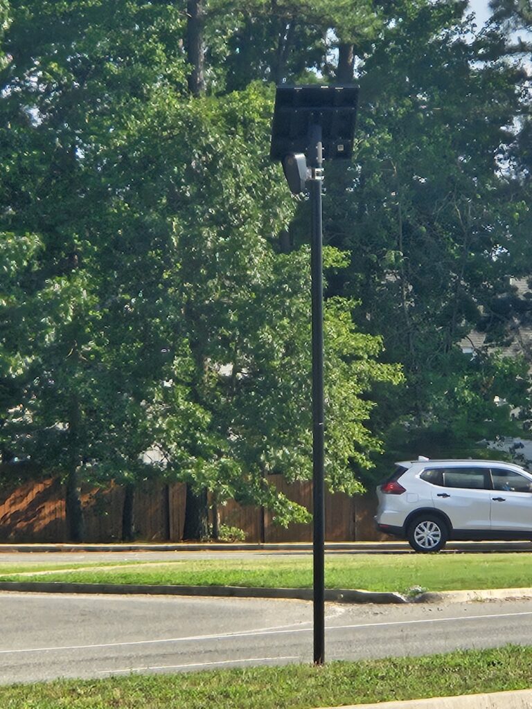 Virginia’s State of Surveillance 1 A surveillance camera stands by a road with a white SUV parked nearby. Green trees and a wooden fence are in the background.